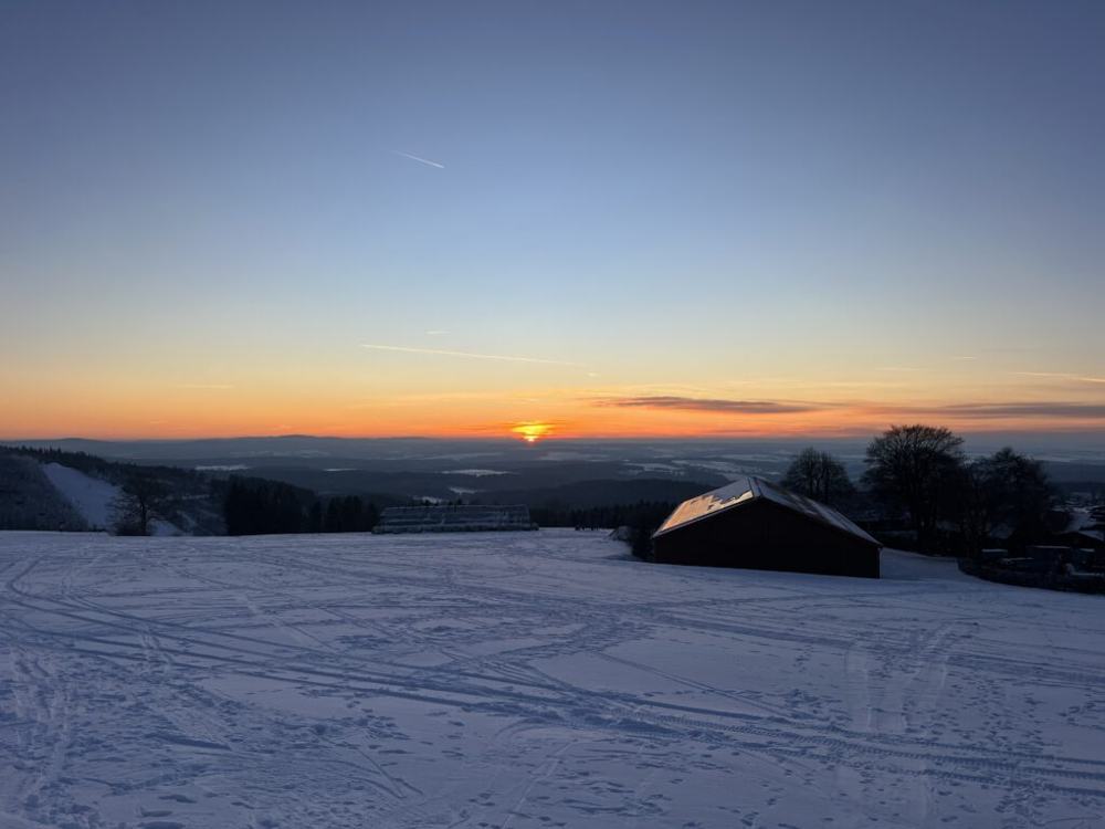 Verschneiter Hang im Abendlicht, im Hintergrund eine Hütte und weite Sicht ins Tal